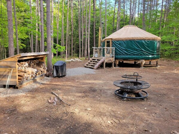 Un terrain de camping rustique doté d’un chalet en bois, d’un foyer et d’un espace de repos couvert, le tout entouré de grands arbres.
