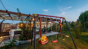 A swing set in a backyard with a patio area, outdoor furniture, and a house in the background. - Villa Marijana (Galovac)