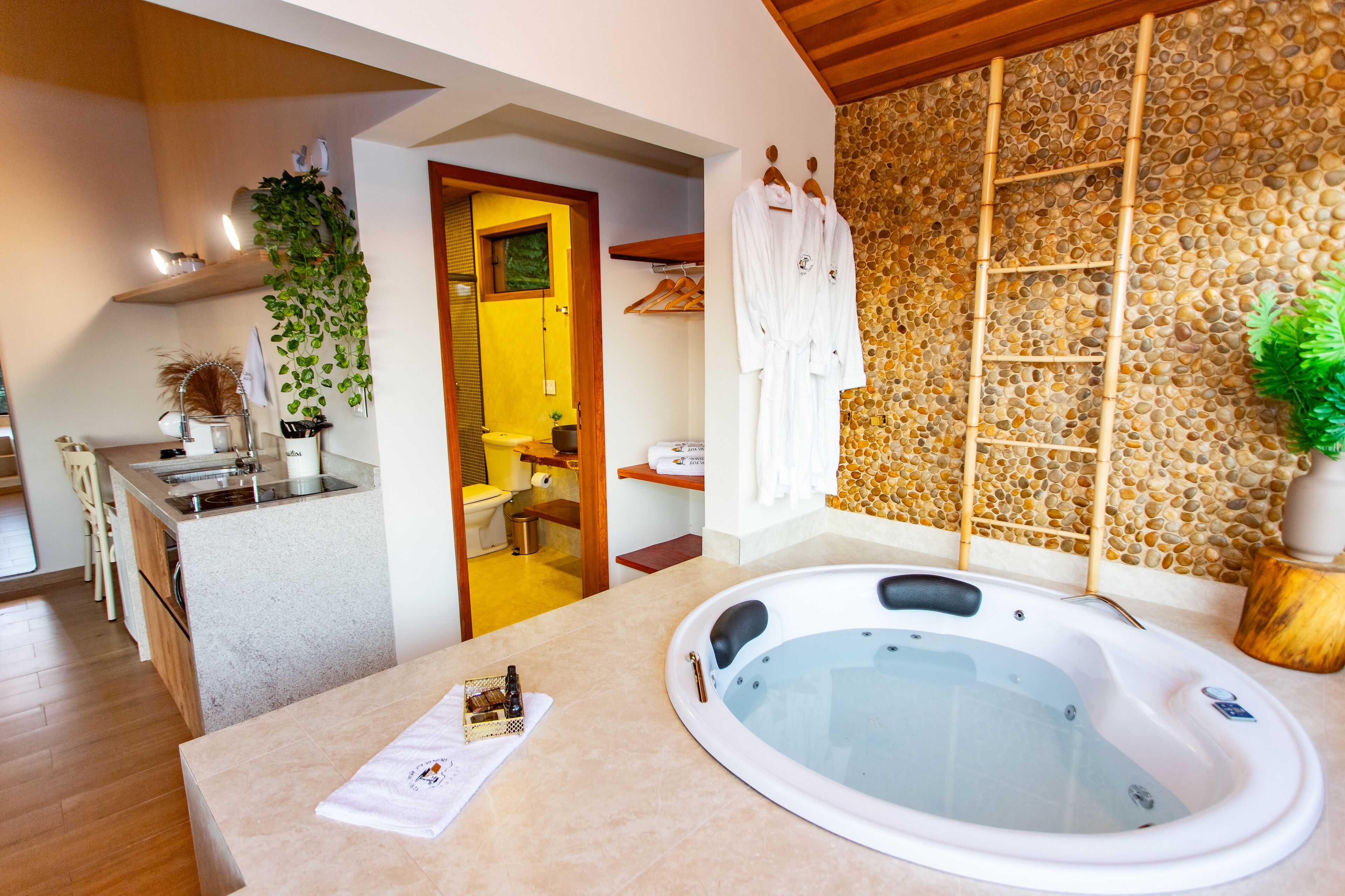 A modern bathroom with a large bathtub, a shower area, and a toilet. There is a wooden shelf with towels and a potted plant.