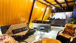 Uma sala de estar moderna com um sofá de couro preto, uma mesa de centro de madeira e uma poltrona revestida de pele. Uma cozinha com geladeira de aço inoxidável e um espaço de bar é visível ao fundo.