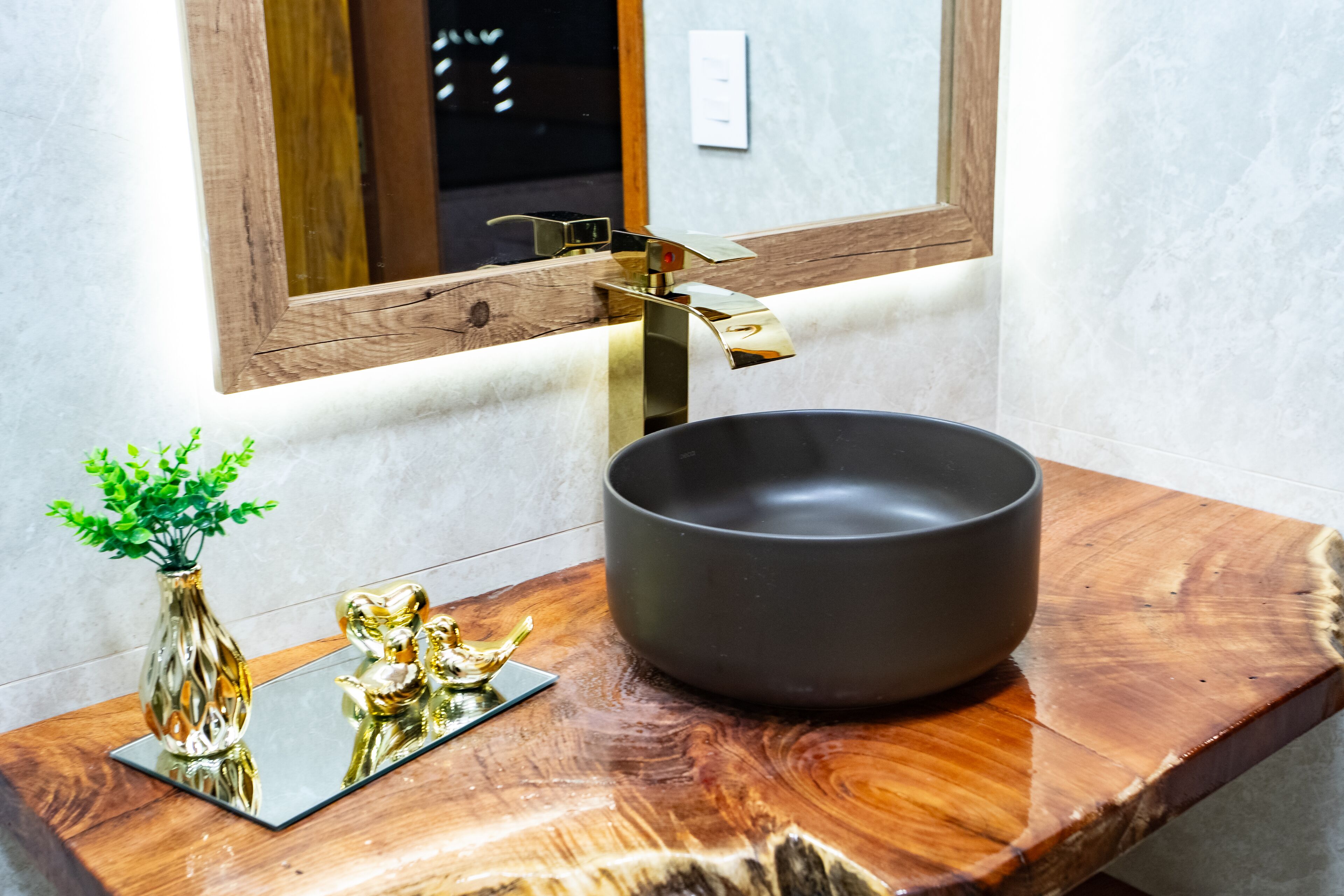 Casa de banho com toucador de madeira, lavatório preto embutido e um objeto decorativo dourado, além de uma pequena planta num vaso.