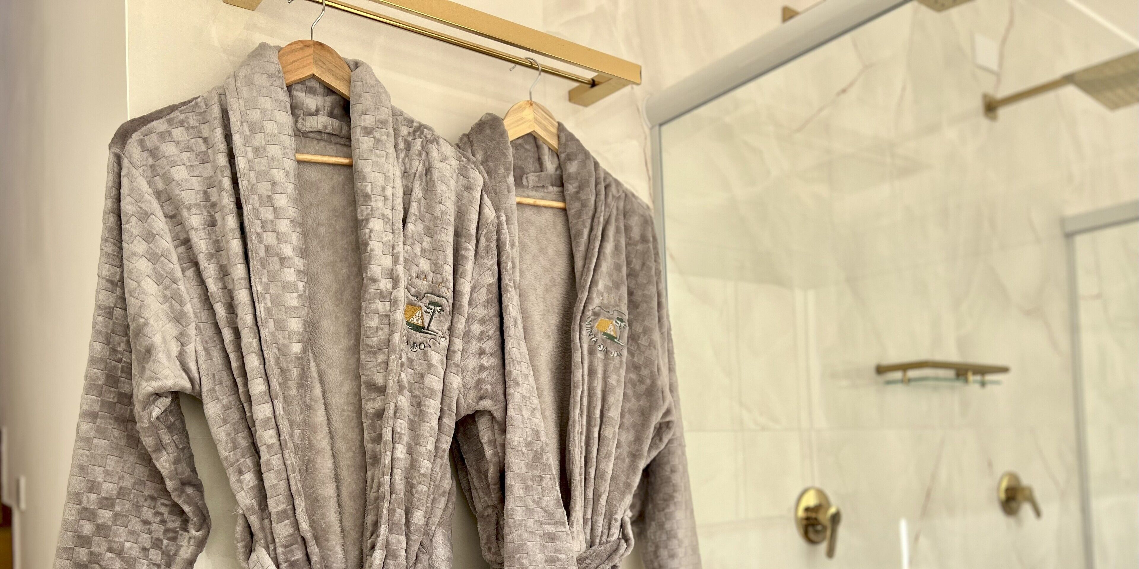 Two grey robes hanging on a wooden rack in a bathroom with a glass shower door.