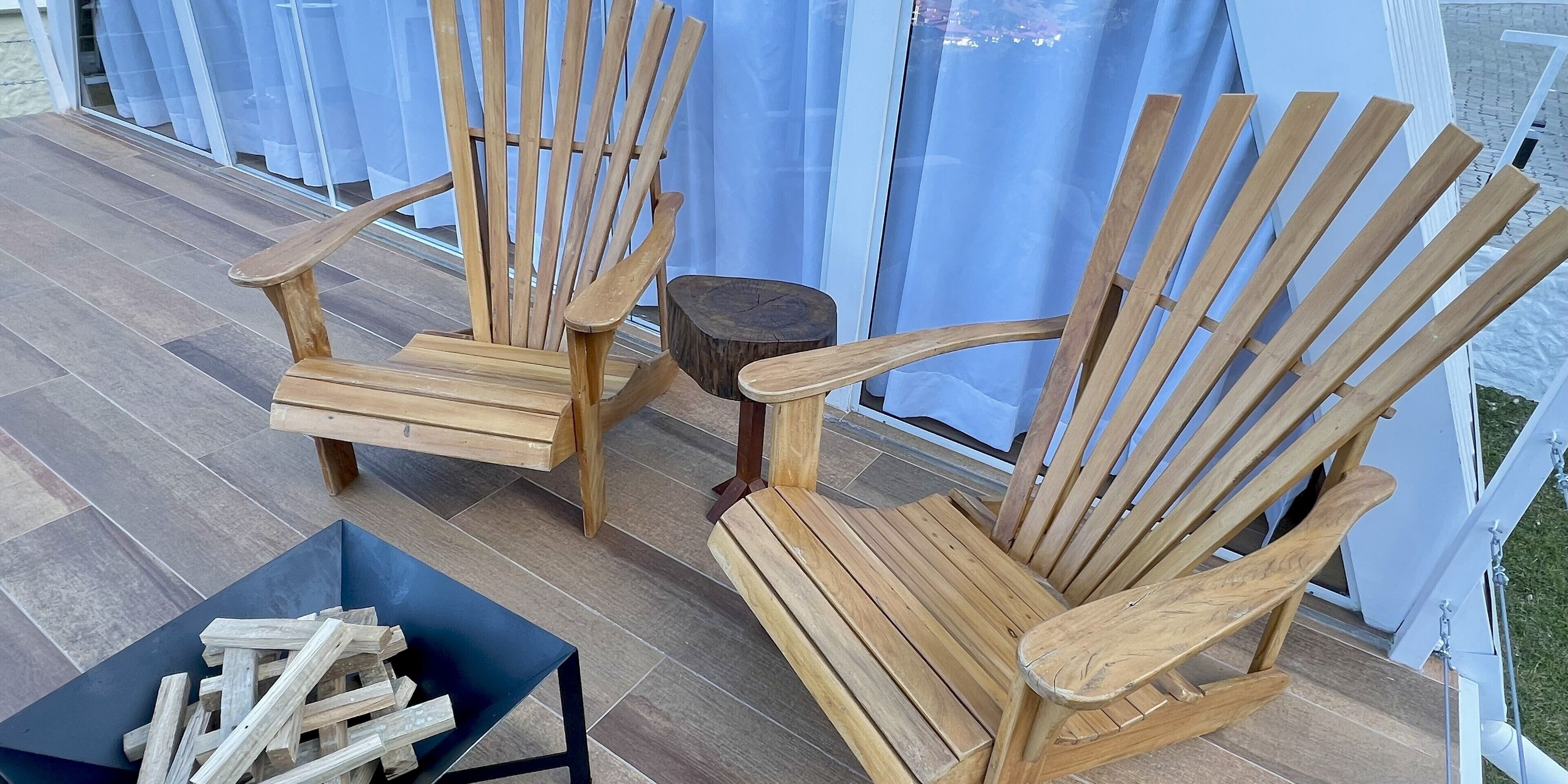 Two wooden Adirondack chairs on a wooden deck with a small table and a black box containing wooden logs.