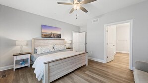 Une chambre à coucher comprenant un lit, des tables de chevet, un ventilateur de plafond, un tableau et une porte donnant sur une autre pièce.
