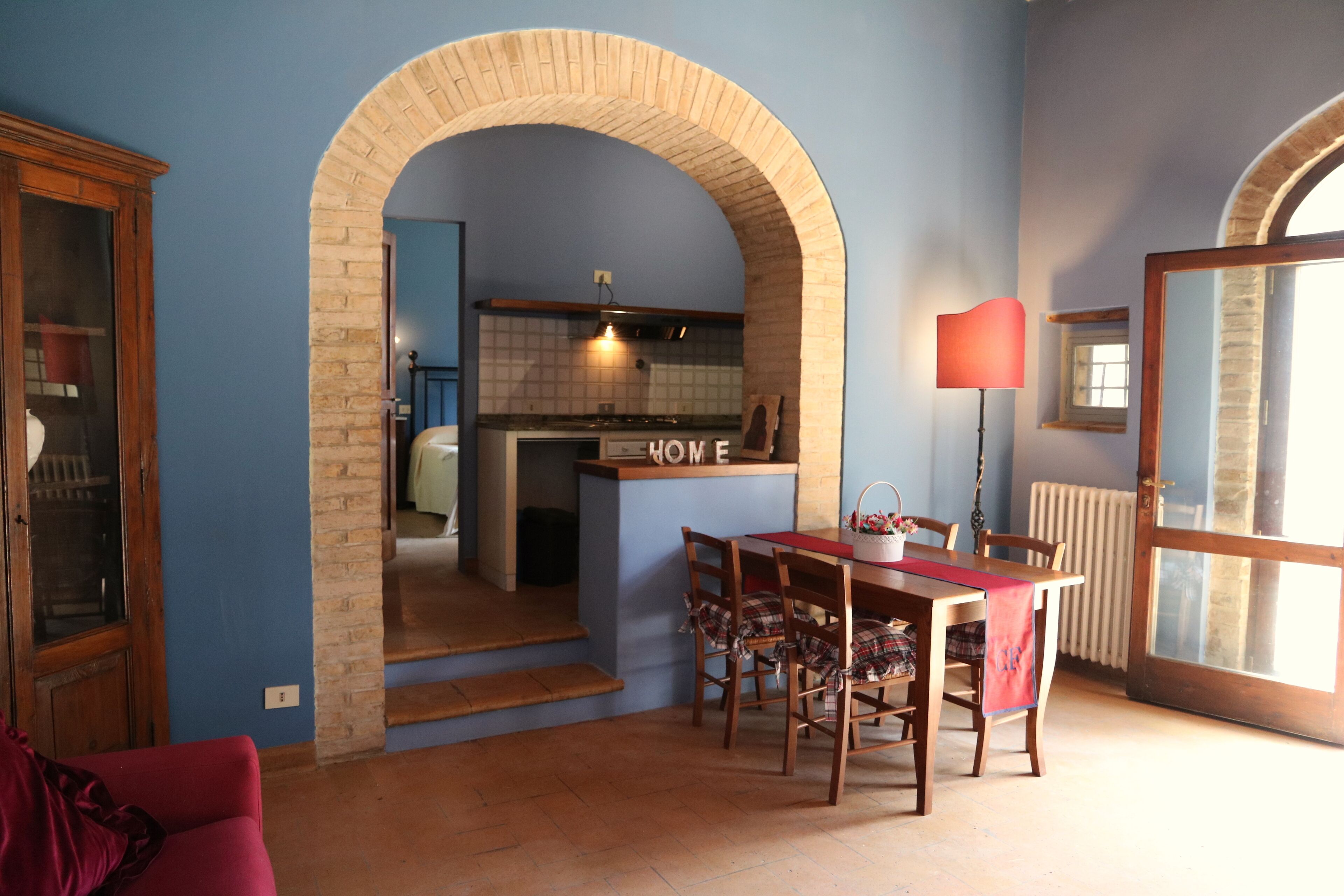 Un'area pranzo con un tavolo e sedie rossi, una cucina visibile attraverso un arco, e una lampada rossa.