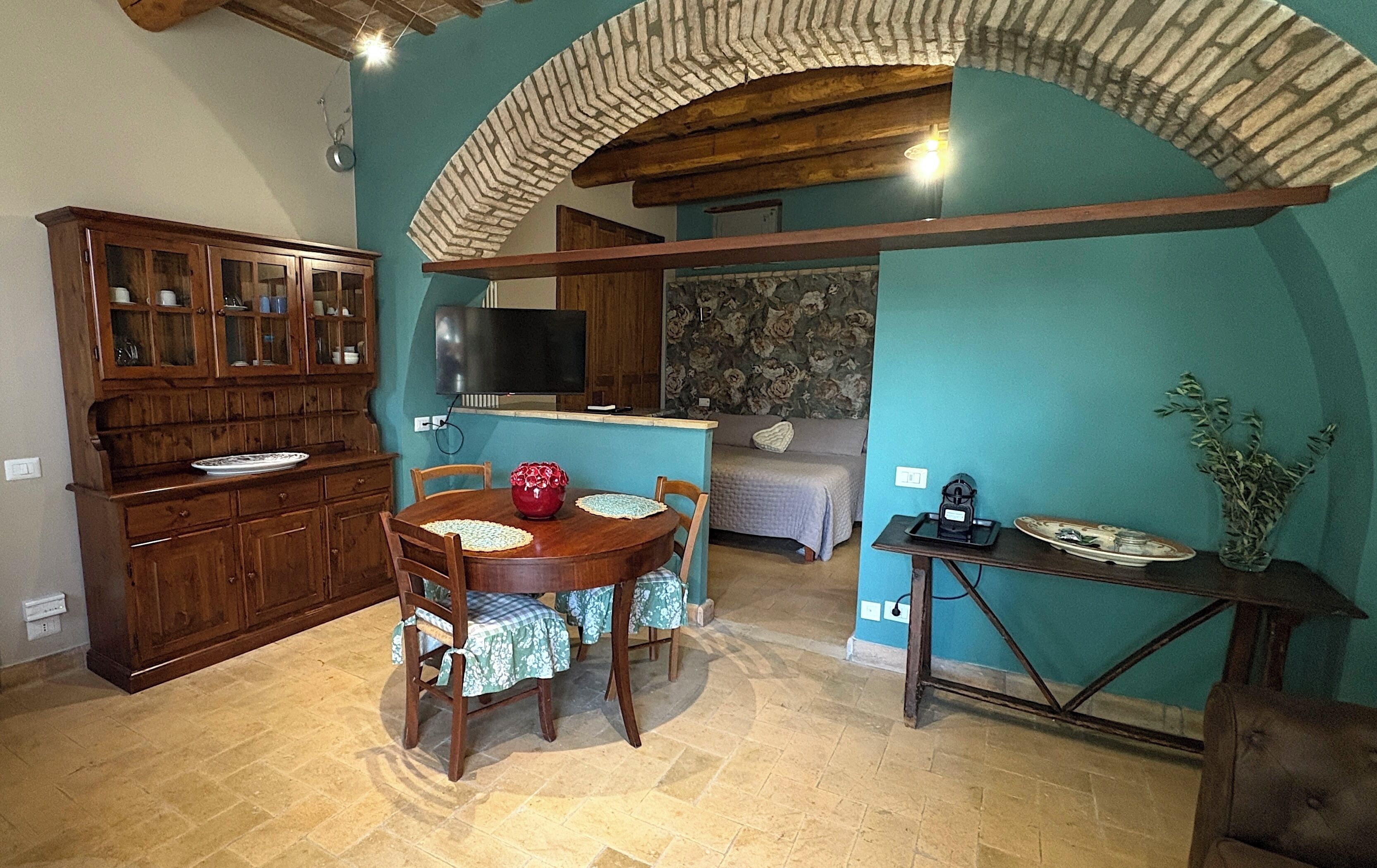 Un comedor con mesa y sillas de madera, una cocina con armarios, un televisor y un sofá al fondo.