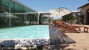 A modern pool area with clear blue water, wooden loungers, and a glass-enclosed lounge area. - Corte Fratini Residenza D'Epoca (Trevi)