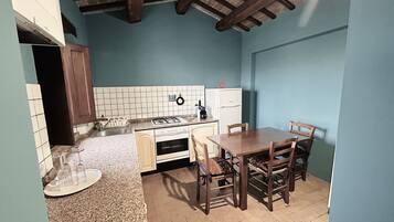 A compact kitchen with a gas stove, sink, and a small refrigerator. There is a dining table with chairs and a tiled backsplash.