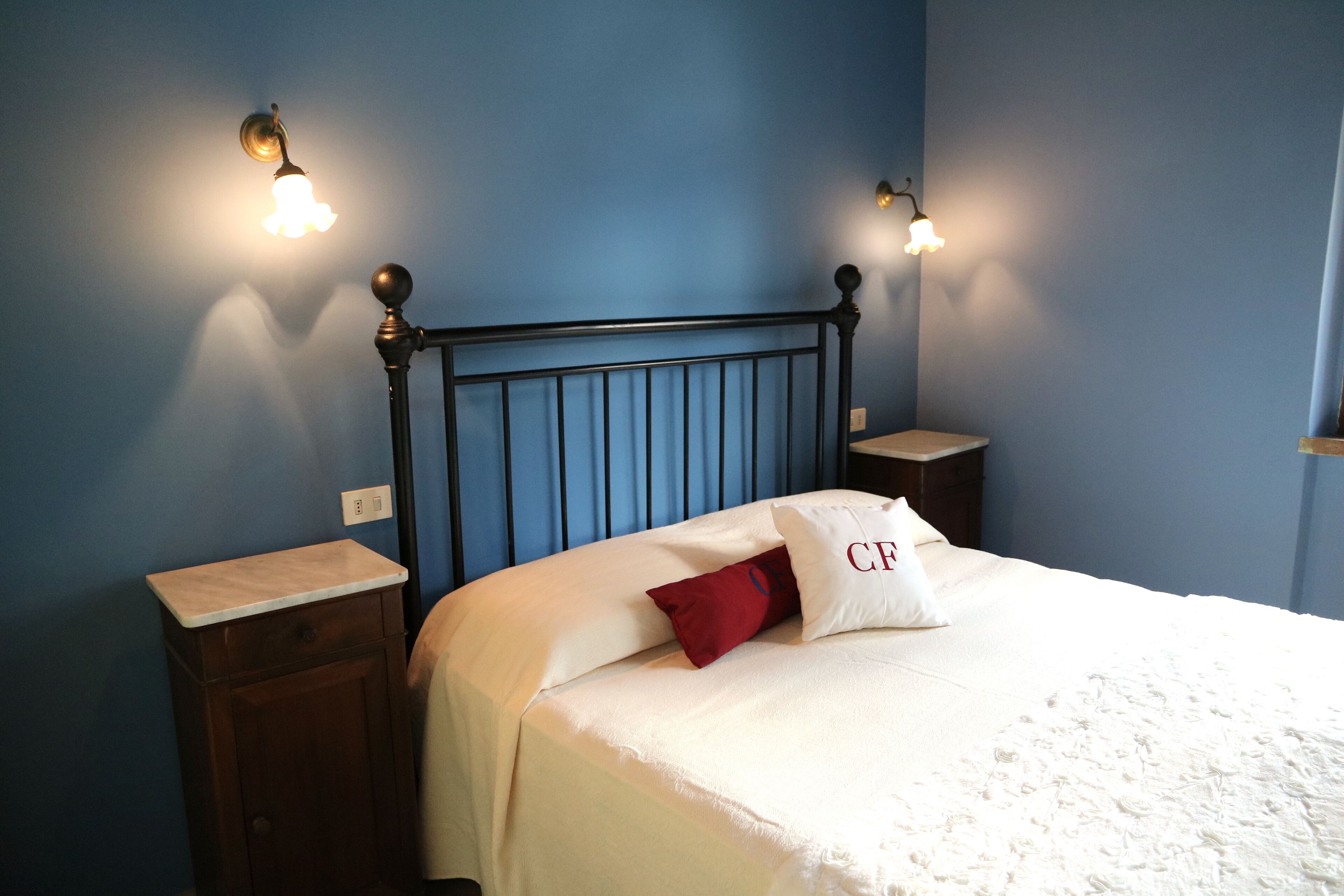 Un dormitorio con pared azul, cabecera de madera, colcha blanca y dos mesitas de noche con lámparas.