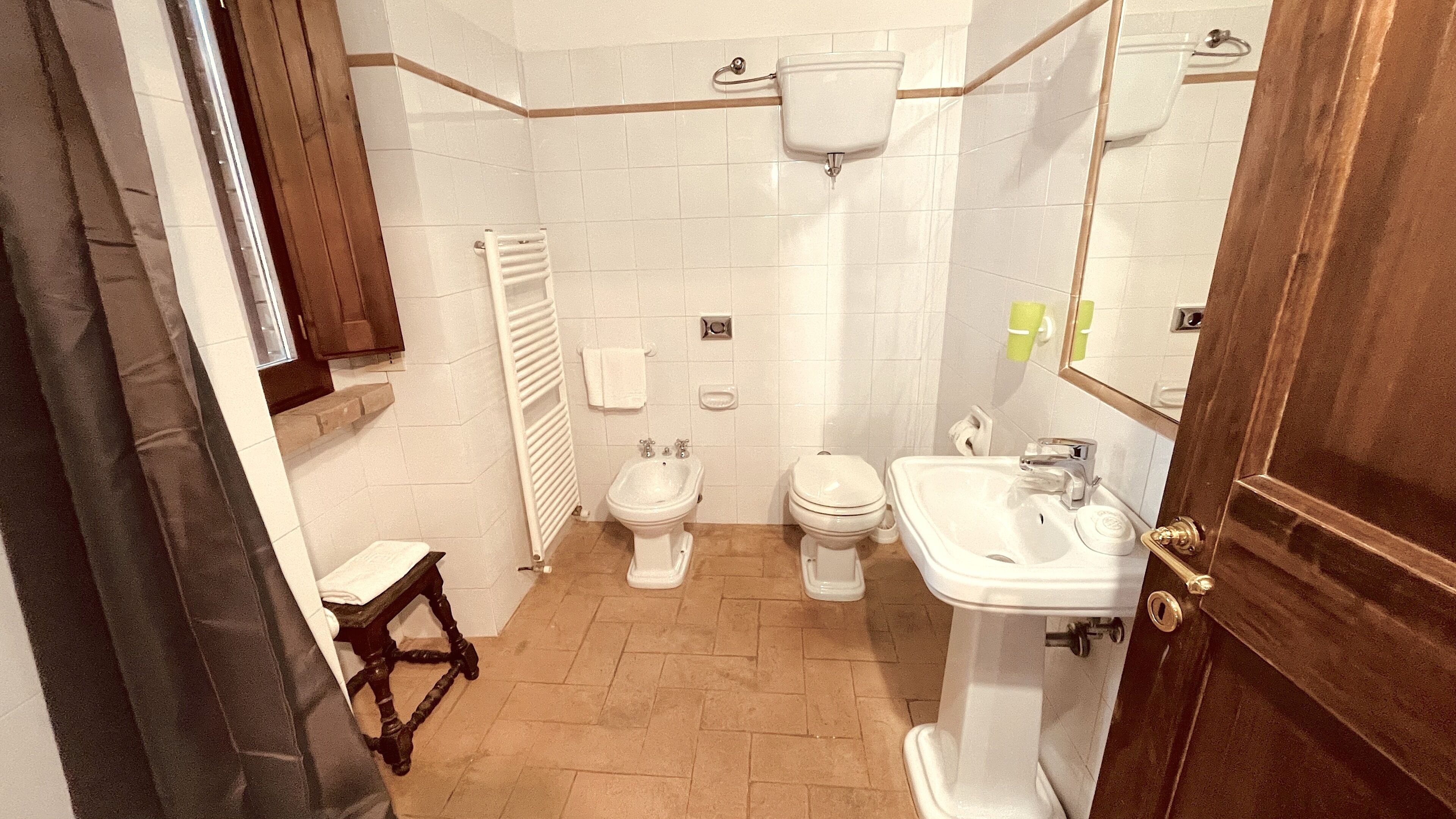 Ein Badezimmer mit Toilette, Bidet und Waschbecken. Ein Handtuchhalter und ein kleiner Hocker sind vorhanden.