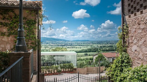 A view from a balcony with a stone wall, a metal railing, and a glass window overlooking a rural landscape with fields and distant hills. - Corte Fratini Residenza D'Epoca (Trevi)
