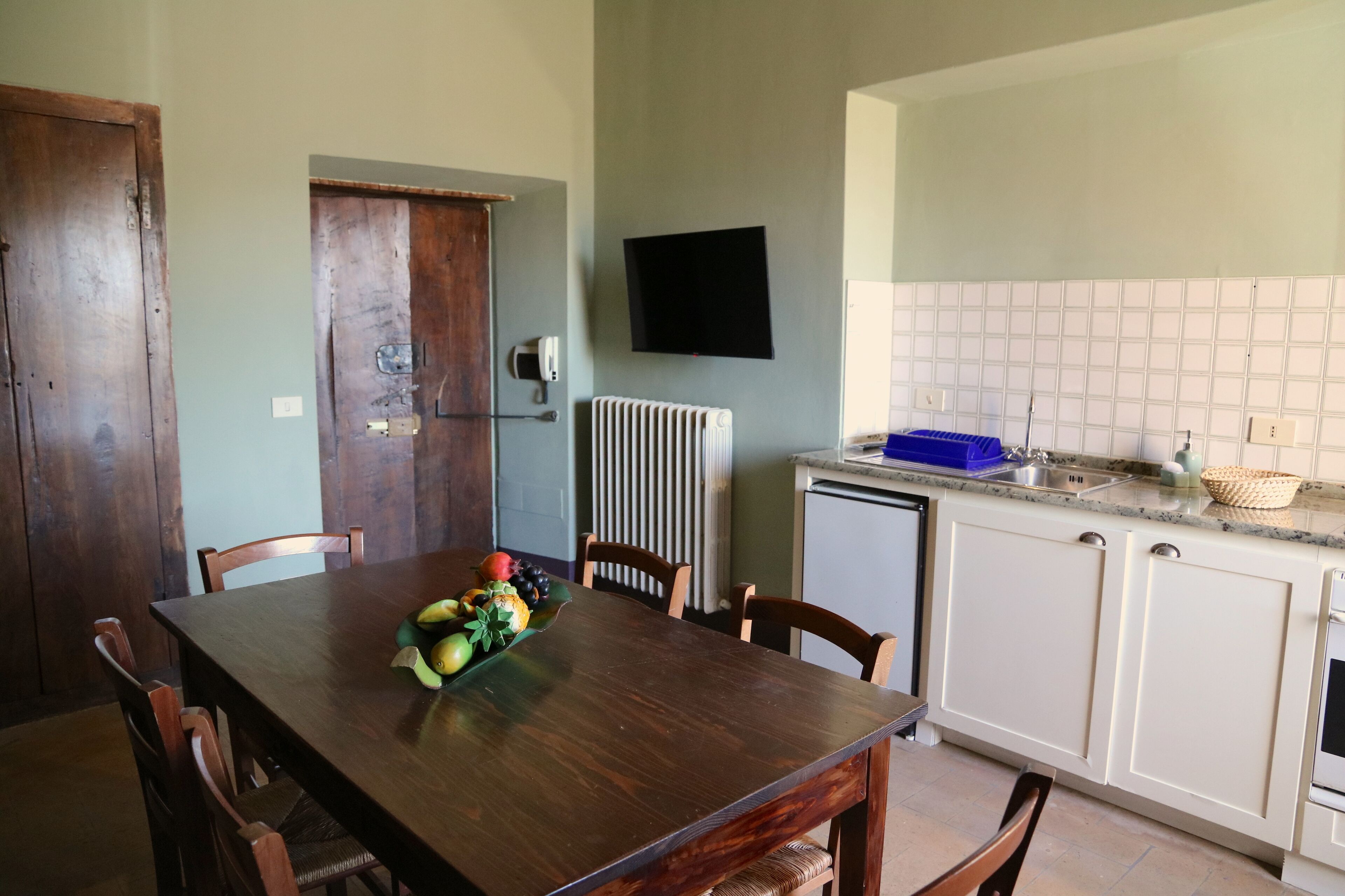 Un coin repas avec une table et des chaises en bois, un panier de fruits, un téléviseur fixé au mur, et une cuisine avec des meubles blancs et un dosseret carrelé.