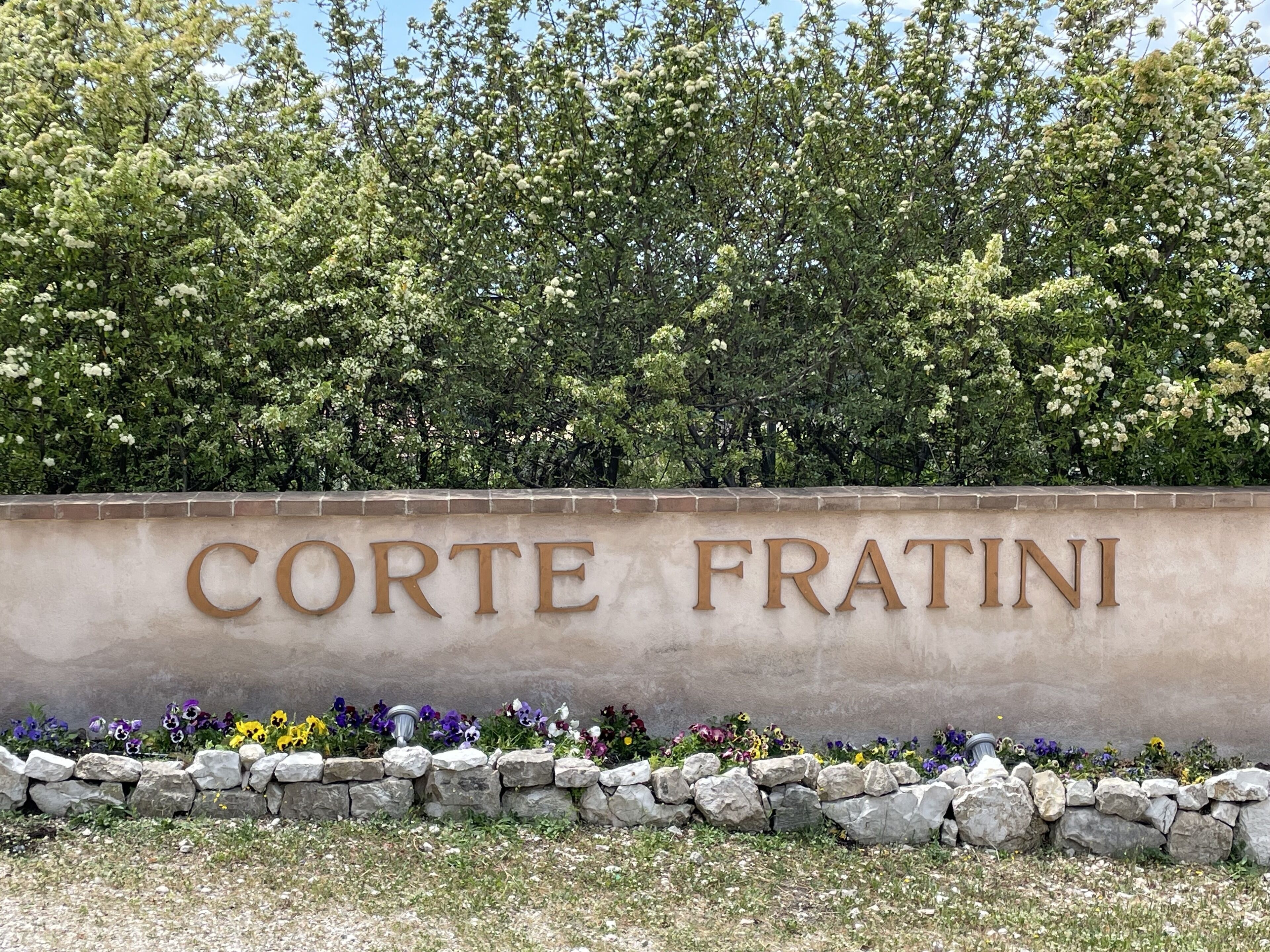 Un letrero con el nombre 'Corte Fratini' rodeado de un muro de piedra y flores coloridas.