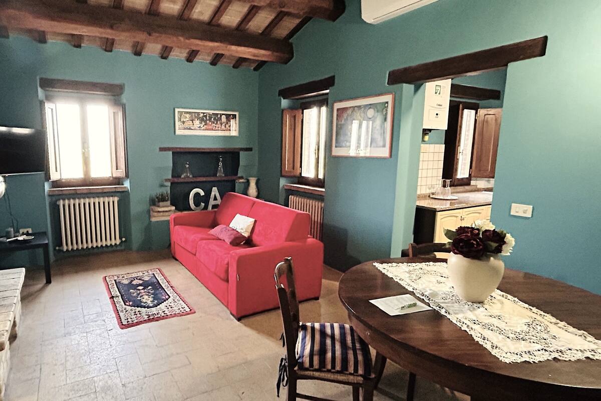 Un salón acogedor con un sofá rojo, una mesa de comedor de madera con mantel de encaje, una silla a rayas, una chimenea y una ventana con vistas al exterior.