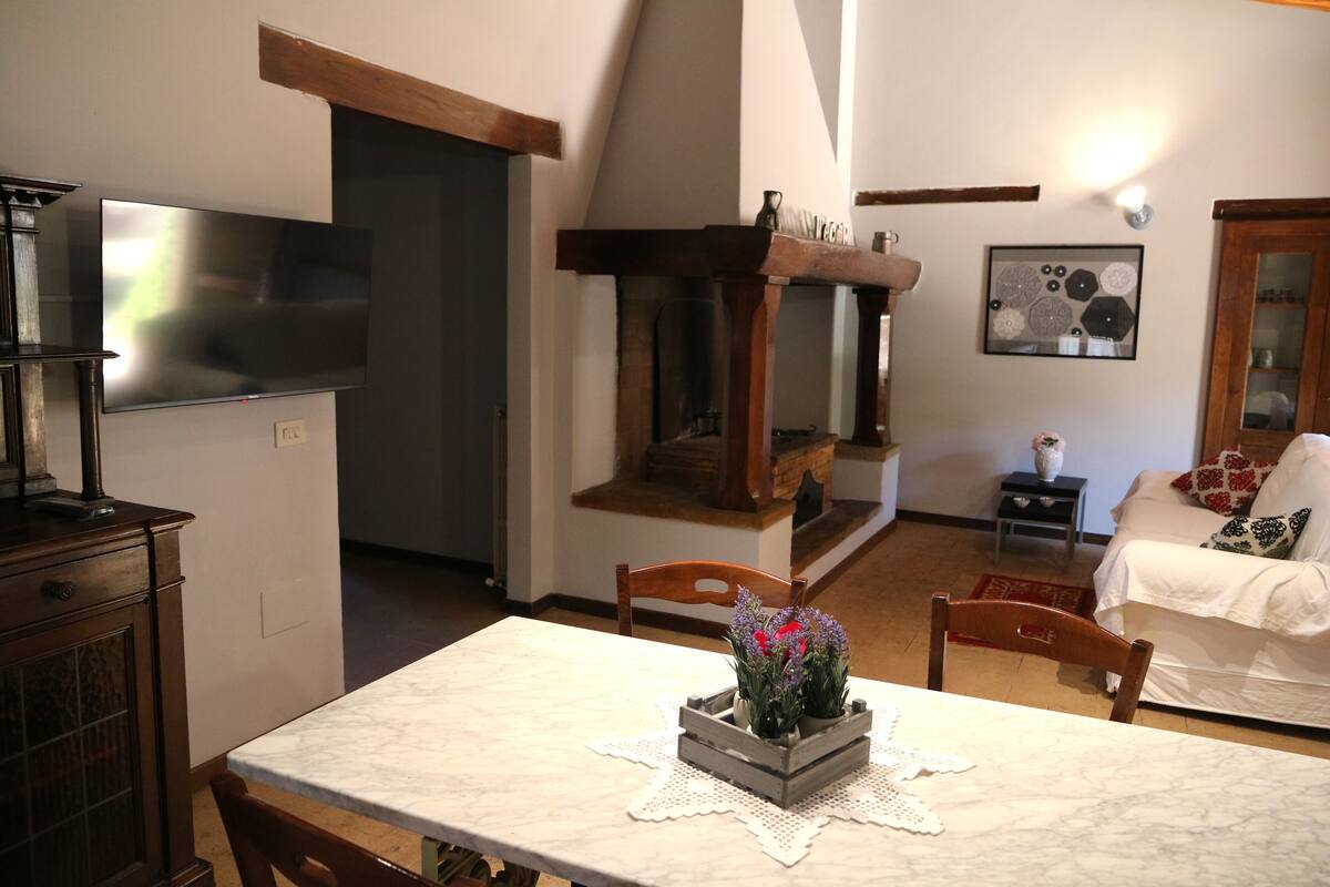 Un comedor con una mesa de mármol, sillas y un centro de mesa floral. Se observa un televisor montado en la pared, una chimenea y un sofá al fondo.
