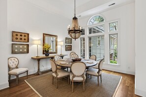 A dining room with a round table, chairs, a chandelier, and large windows. - Luxury 4BD on Kiawah w/ Chef's Kitchen, Private Pool + Hot Tub (Kiawah Island)