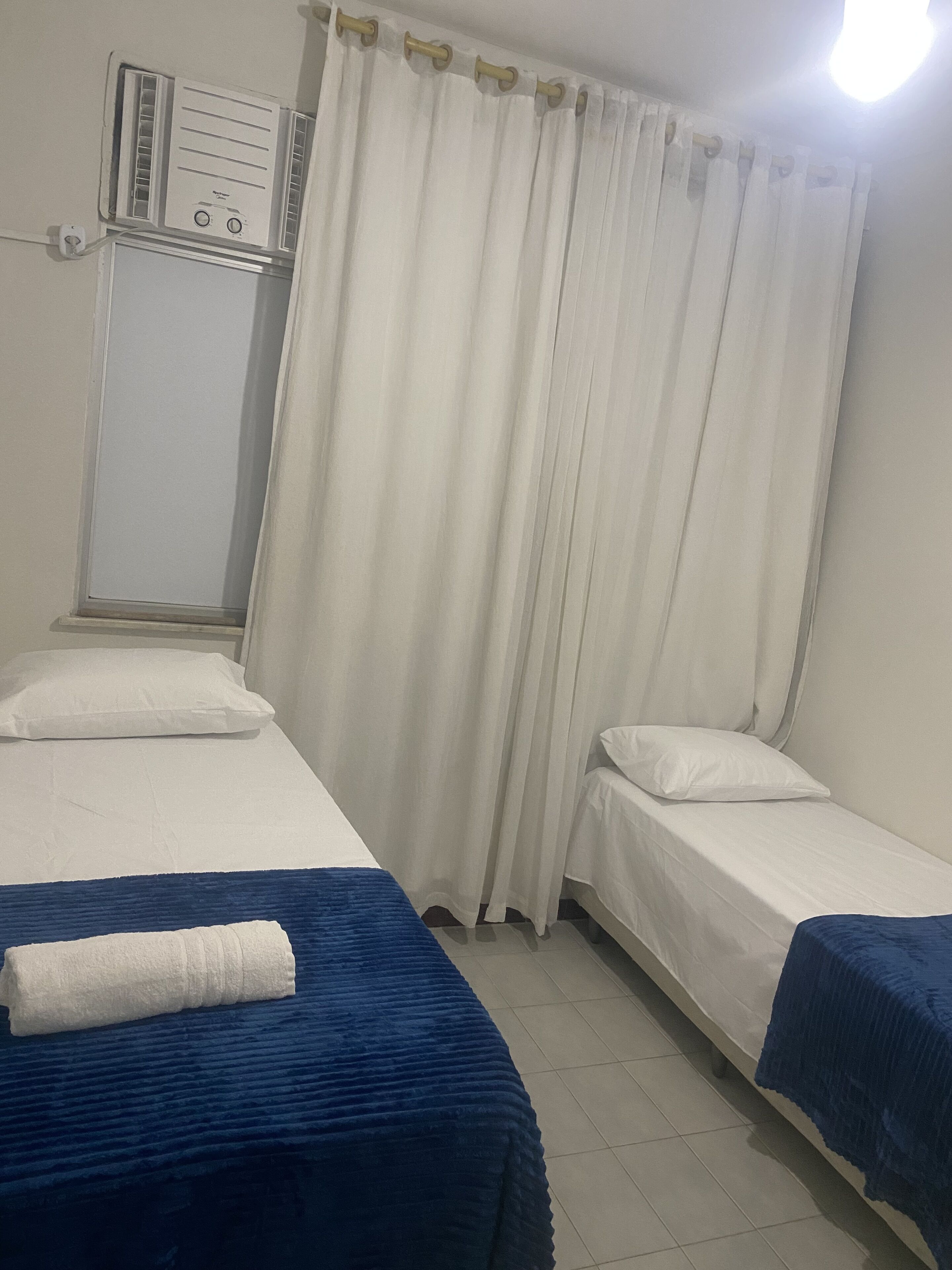 Ein Zimmer mit zwei Betten, einem weißen Vorhang, einem Fenster mit Jalousien und einer Wand-Klimaanlage.