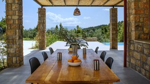 A wooden dining table with chairs, a bowl of oranges, and striped mugs, set under a thatched roof with stone pillars, overlooking a pool and lush greenery. - The Mavrolitharo Residence, new stone built villa.  (Chalkidiki)
