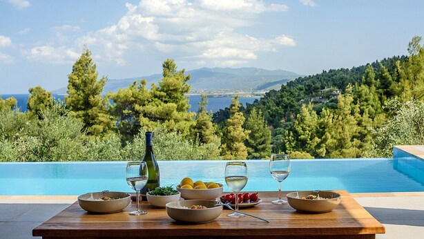 A dining table with bowls of food and wine glasses, overlooking a pool and a scenic view of trees and mountains. - The Mavrolitharo Residence, new stone built villa.  (Chalkidiki)