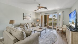 A living room with a beige sofa, a coffee table, and a TV. There is a ceiling fan, a lamp, and a painting on the wall. - Completely Remodeled, 2 Kings, Bunkroom, Beach Service  (Miramar Beach)