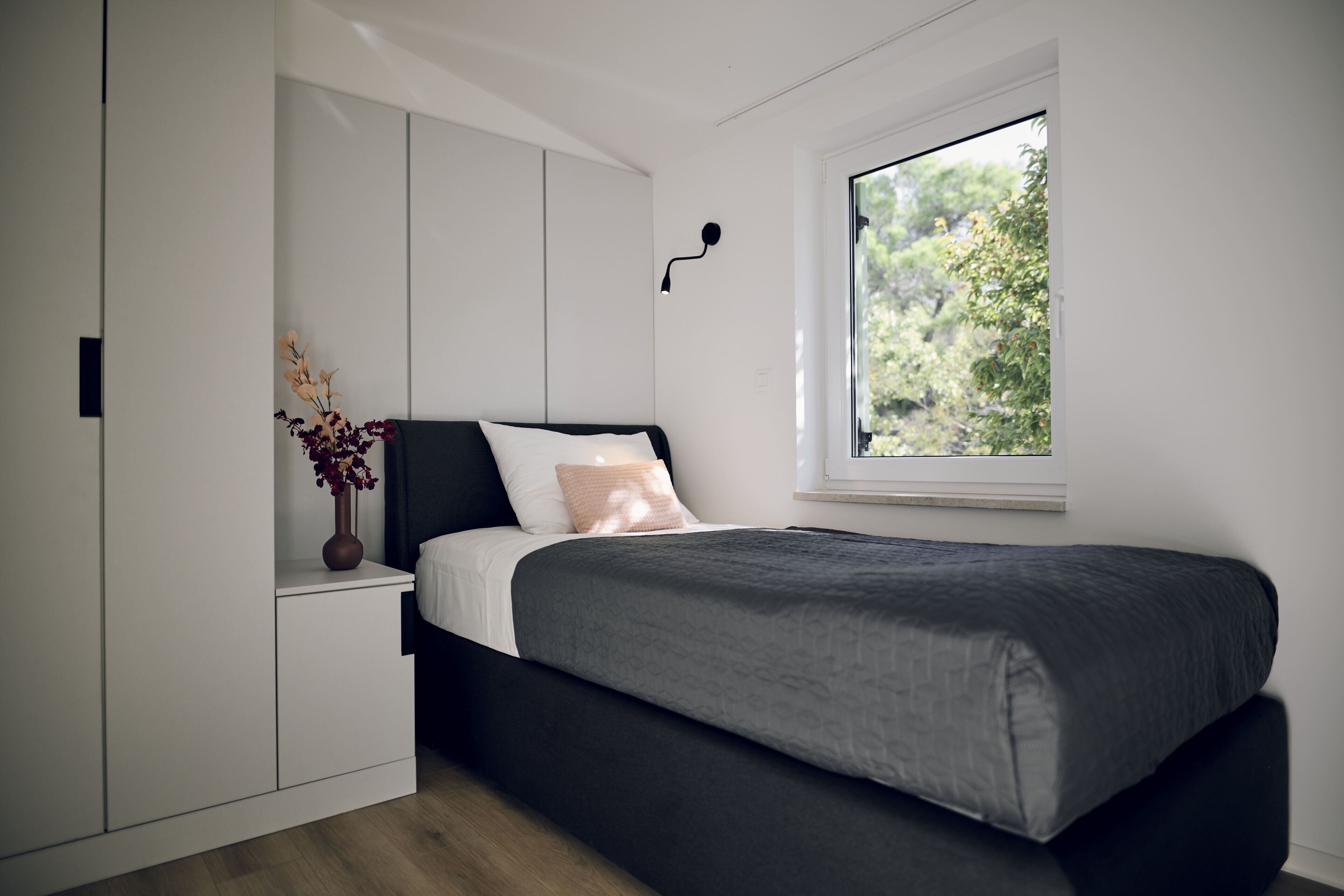 A modern bedroom with a bed, built-in wardrobe, and a window with a view of greenery.