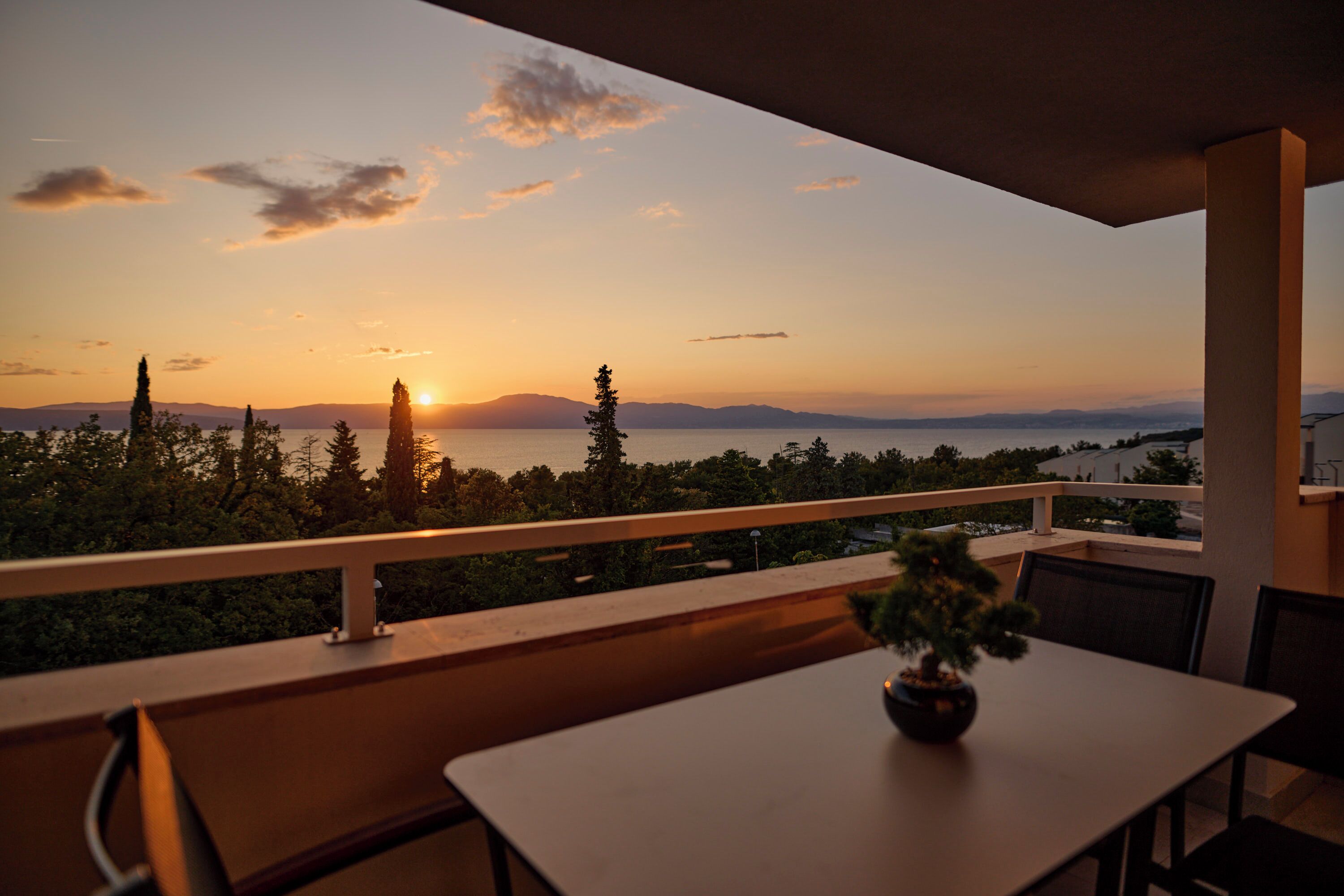 En balkon med et bord og stole, der giver udsigt til solnedgang over træer og fjerne bjerge.