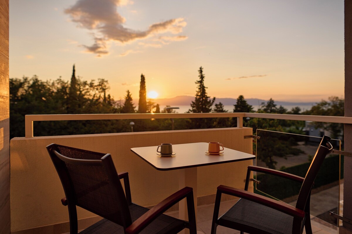 Un balcon avec deux chaises et une table, d’où l’on peut admirer un coucher de soleil sur un paysage arboré et une montagne lointaine.