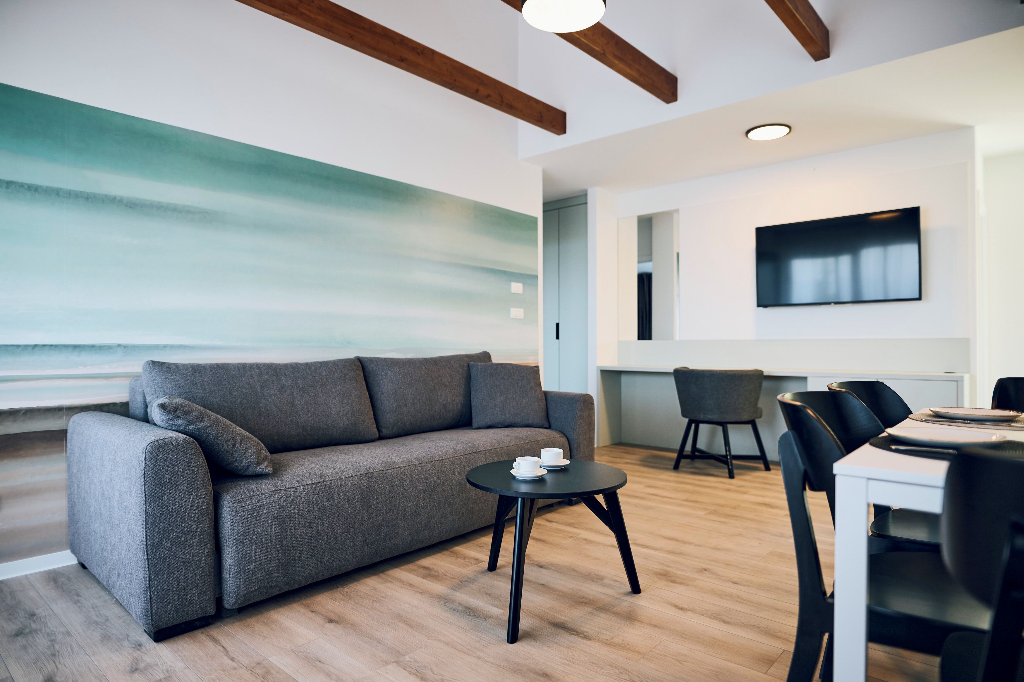 Een moderne woonkamer met een grijze bank, een klein rond tafeltje met een kop en schotel, een eethoek met een witte tafel en zwarte stoelen, en een groot abstract schilderij aan de muur.