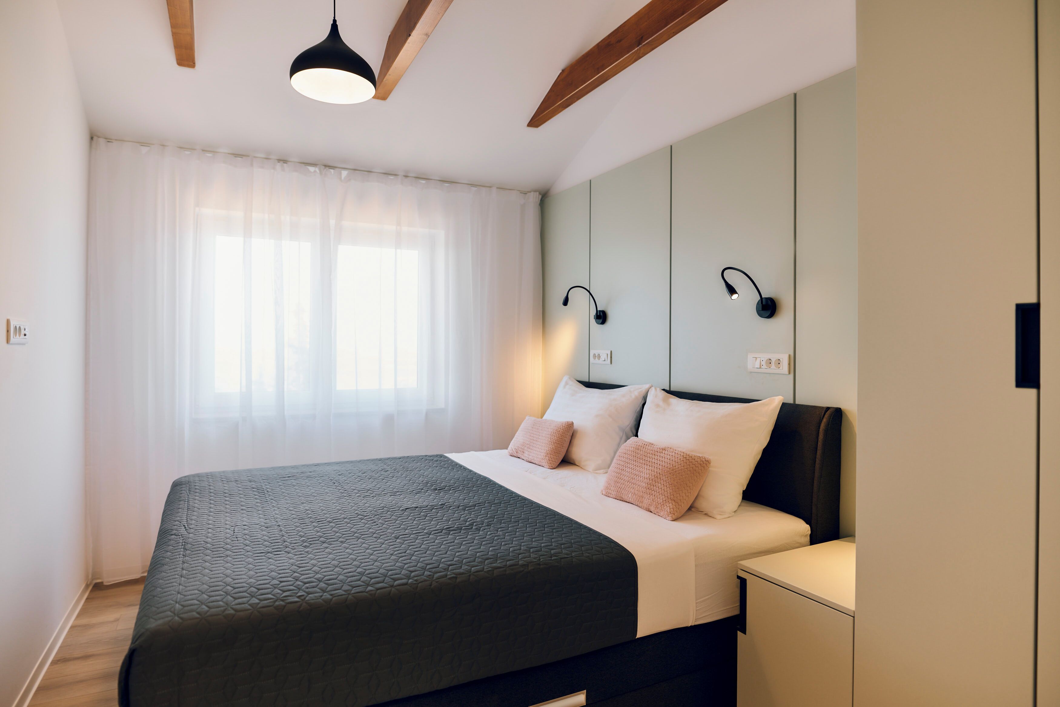 Een moderne slaapkamer met een groot bed, een nachtkastje, een ventilator in het plafond en een raam met dunne gordijnen.