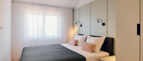 Een moderne slaapkamer met een groot bed, een nachtkastje, een ventilator in het plafond en een raam met dunne gordijnen.