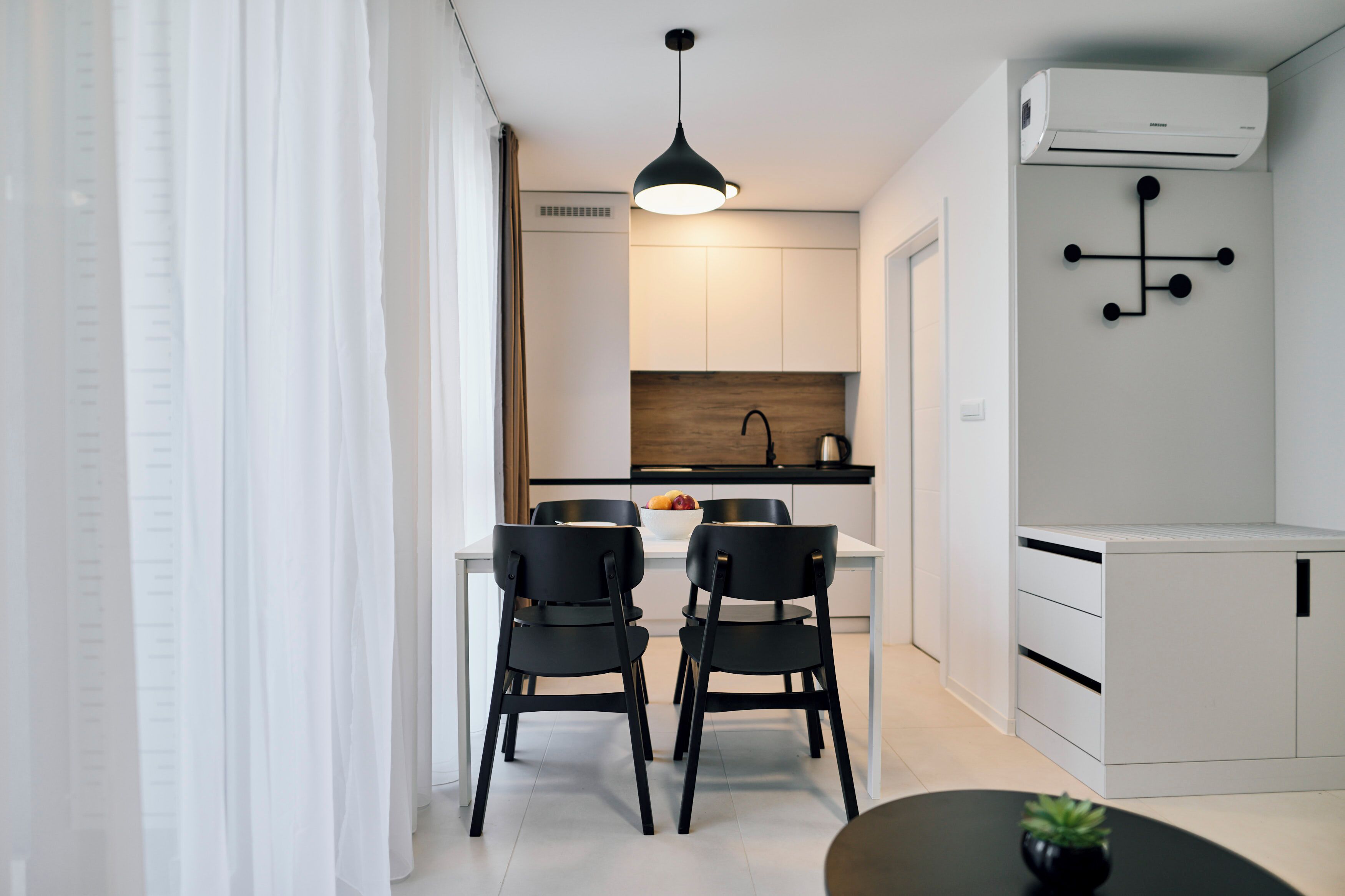 Une cuisine moderne avec des meubles blancs, un luminaire suspendu noir et un petit coin repas agrémenté de deux chaises noires.