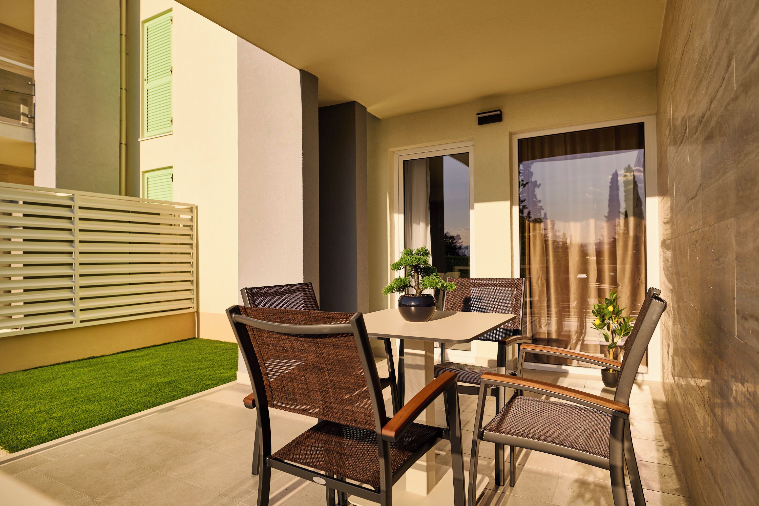 Uma área de refeições ao ar livre com mesa e cadeiras, um pequeno vaso com planta e vista para um edifício com persianas verdes.