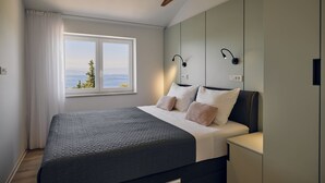 A bedroom with a bed, a nightstand, a window with curtains, and a view of the outdoors. - Malinska Green Apartments (Malinska-Dubasnica)