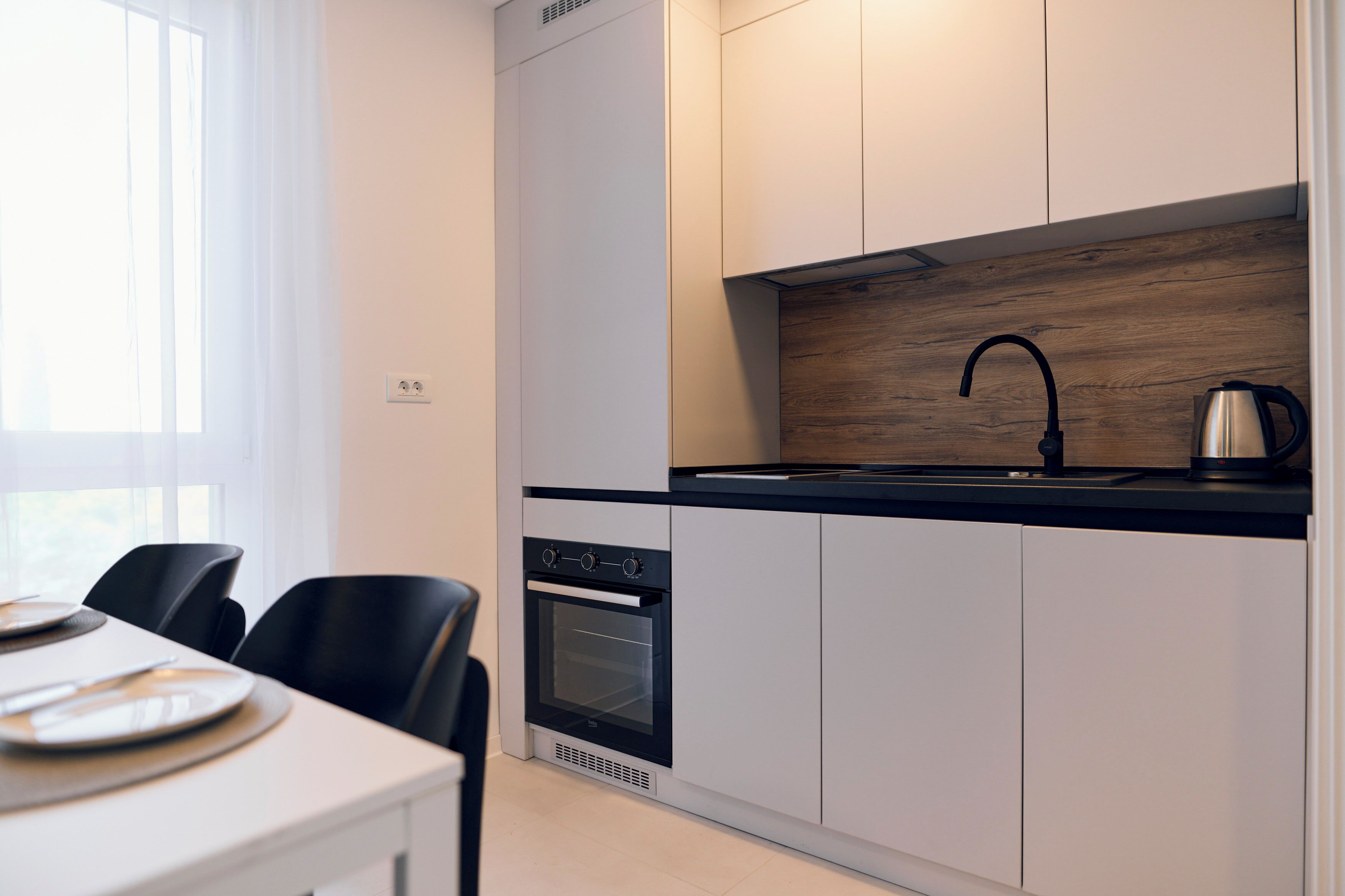 A modern kitchen with white cabinets, a black countertop, a built-in oven, and a kettle.