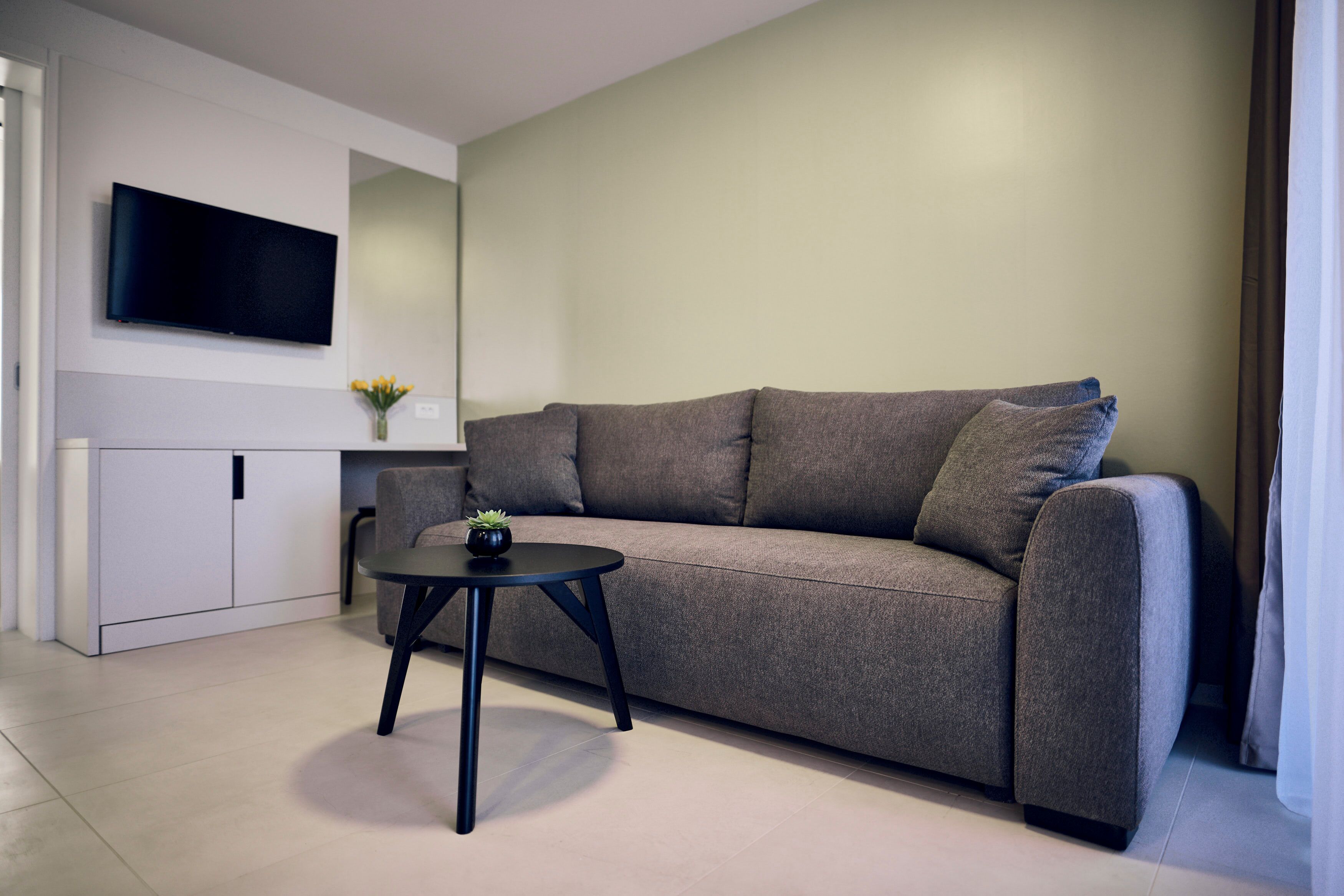 Een moderne woonkamer met een grijze bank, een klein zwart tafeltje en een flatscreen televisie die aan de muur is bevestigd.
