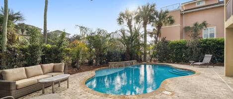 Una piscina de borde curvo, rodeada por un patio de ladrillo. Hay un sofá color beige y una pequeña mesa con sillas.