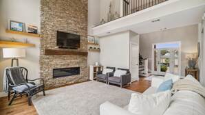 Un salon spacieux avec une cheminée en pierre, un canapé blanc, un fauteuil gris et un téléviseur à écran plat fixé au mur.