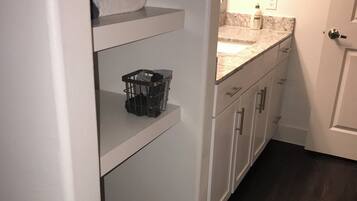 A bathroom with a toilet paper holder, a towel rack with folded towels, a basket, and a countertop with a soap dispenser.