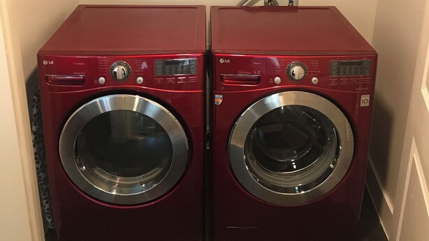 A laundry room with two red LG front-loading washing machines, a shelf above containing cleaning supplies, and a power outlet. - Cozy and Comfortable Legacy West Apartment (Plano)