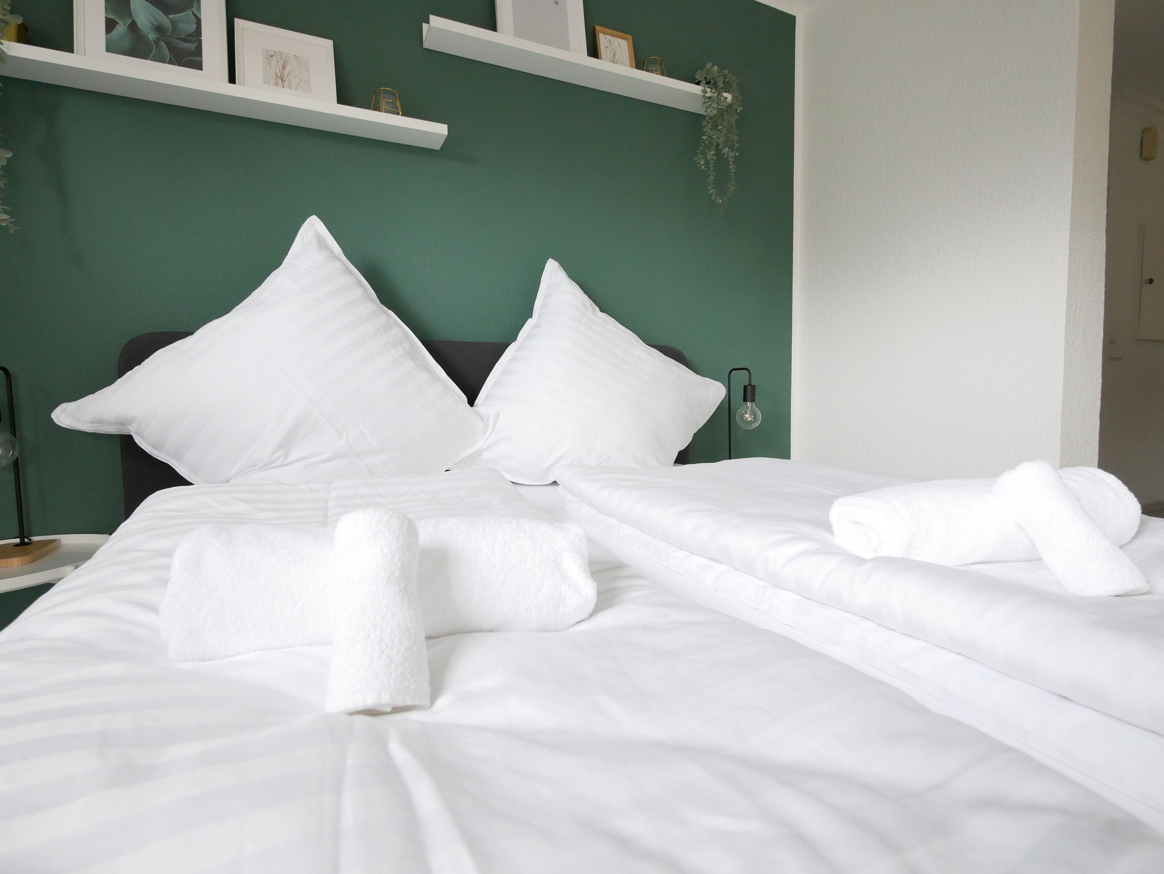 A neatly made bed with white linens and pillows, a bedside lamp, and a wall-mounted shelf with decorative items.