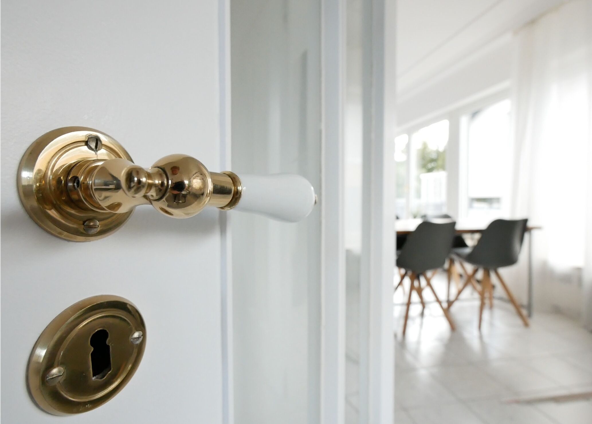 A brass door handle and lock, a white door, and a view into a room with a dining table and chairs.