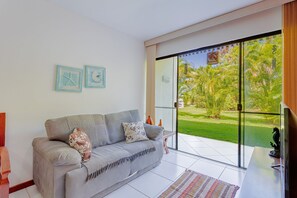 Een moderne woonkamer met een grijze bank, twee kunstwerken aan de muur, een glazen deur naar een tuin, en een flatscreen televisie op een standaard.