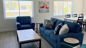 Una sala de estar con un sofá azul, una mesa de centro, sillones verde azulado y una mesa de comedor con sillas, además de un ventilador de techo.