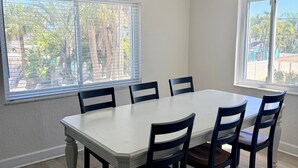 Comedor con mesa blanca y sillas oscuras, ventanas amplias con persianas y un ventilador de techo.