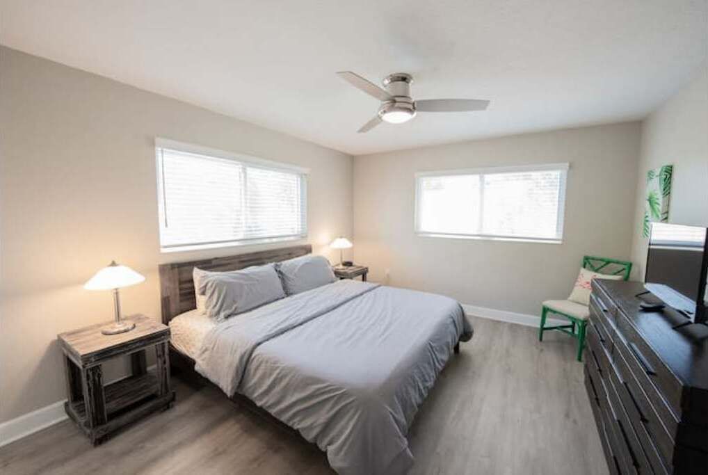 Une chambre à coucher avec un lit, deux tables de chevet, un ventilateur de plafond et deux fenêtres.