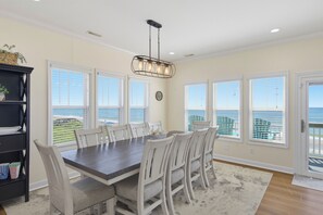 A dining room with a view of the ocean, featuring a large table, white chairs, and a chandelier.