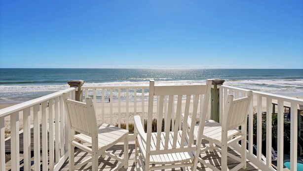 A wooden deck with white chairs overlooking a beach.