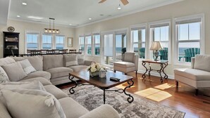 A spacious living room with a large sectional sofa, a coffee table, and a ceiling fan. There are multiple windows providing natural light and views of the outdoors.