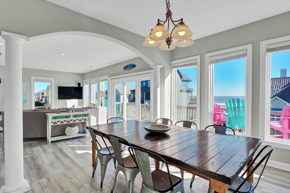 Une salle à manger spacieuse, avec une table et des chaises en bois, un lustre et de grandes fenêtres offrant une vue sur la mer.