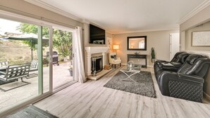 Ein Wohnzimmer mit Kamin, einer Glastür zu einer Terrassenförmigen Außenausstattung, einem schwarzen Ledersofa und einem Glastisch.